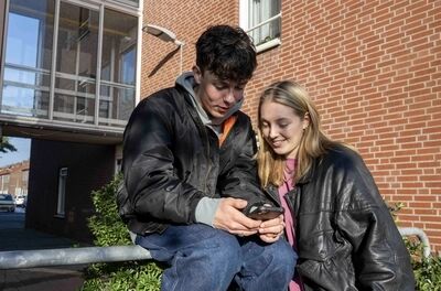 Twee jongeren zitten samen op een railing en kijken naar een smartphone, met een bakstenen gebouw op de achtergrond.